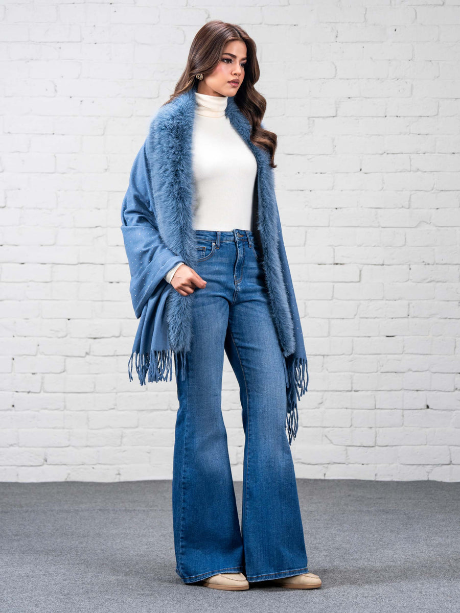 Side view of woman in sky blue wool cape with faux fur trim and fringe, paired with white turtleneck and blue flared jeans