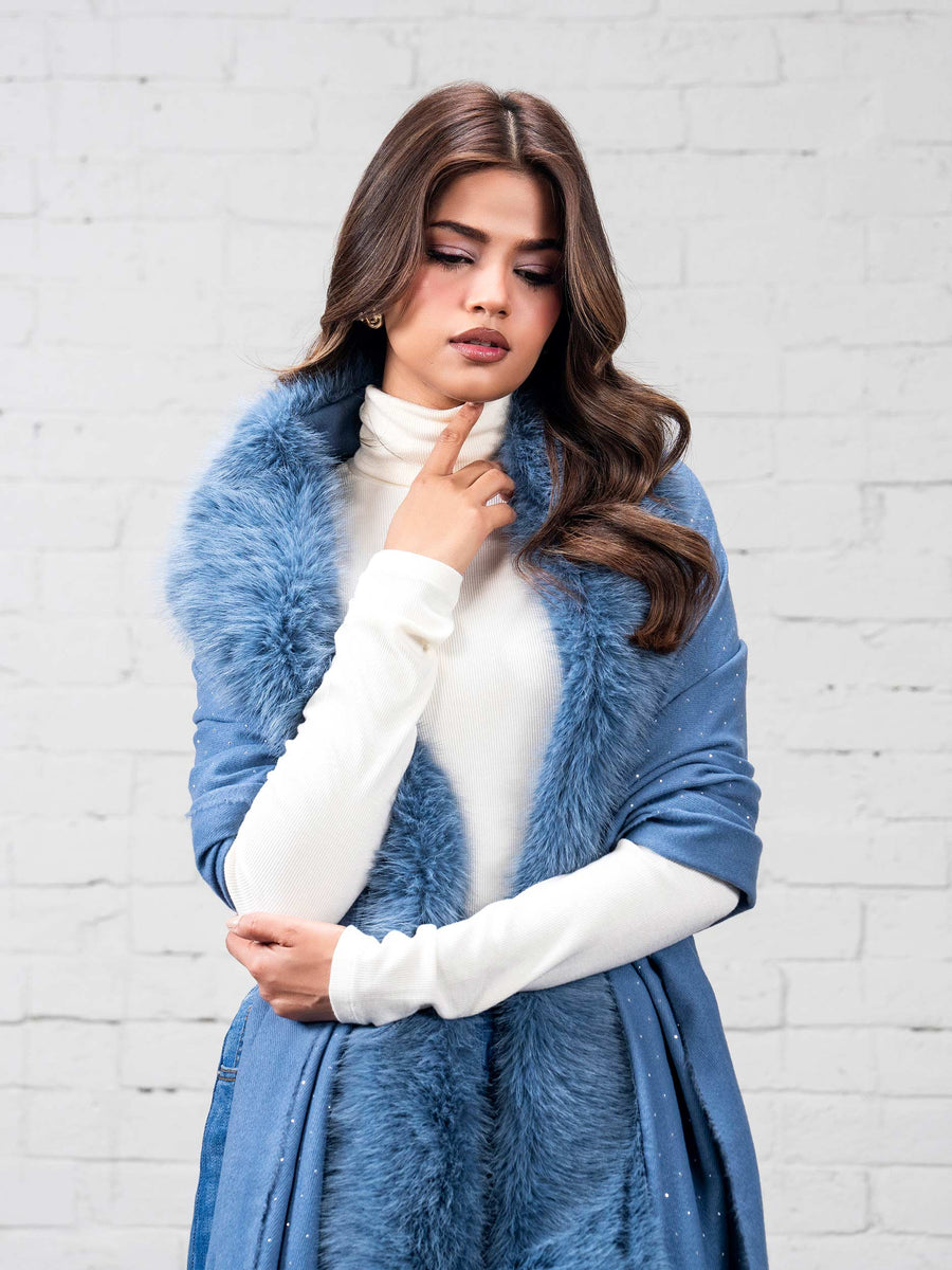 Close-up view of woman thoughtfully posing with hand near chin, wearing sky blue wool cape with faux fur trim and subtle rhinestone details