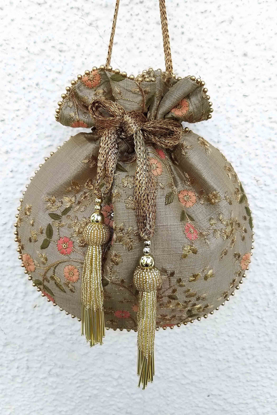 Round embroidered potli bag in grey with peach floral patterns and gold beaded tassels hanging against a white textured wall.