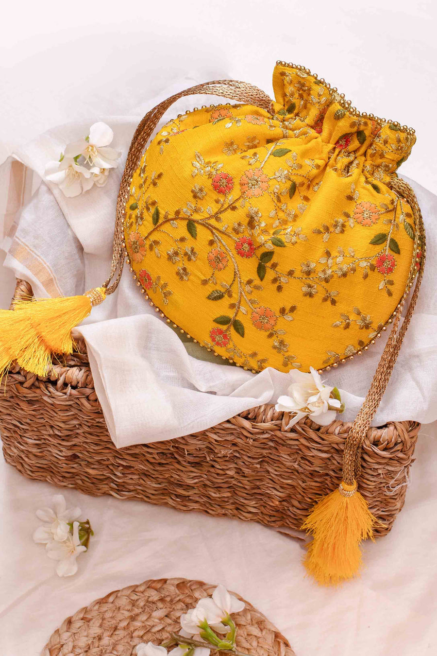 Mustard yellow embroidered floral potli bag with golden drawstring and tassels placed on a woven basket with white flowers.