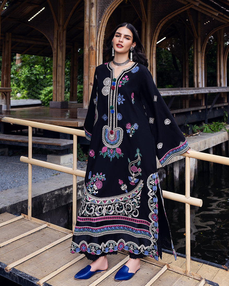 Woman wearing a black Karandi suit with colorful floral embroidery, standing by a bamboo railing with a wooden structure in the background