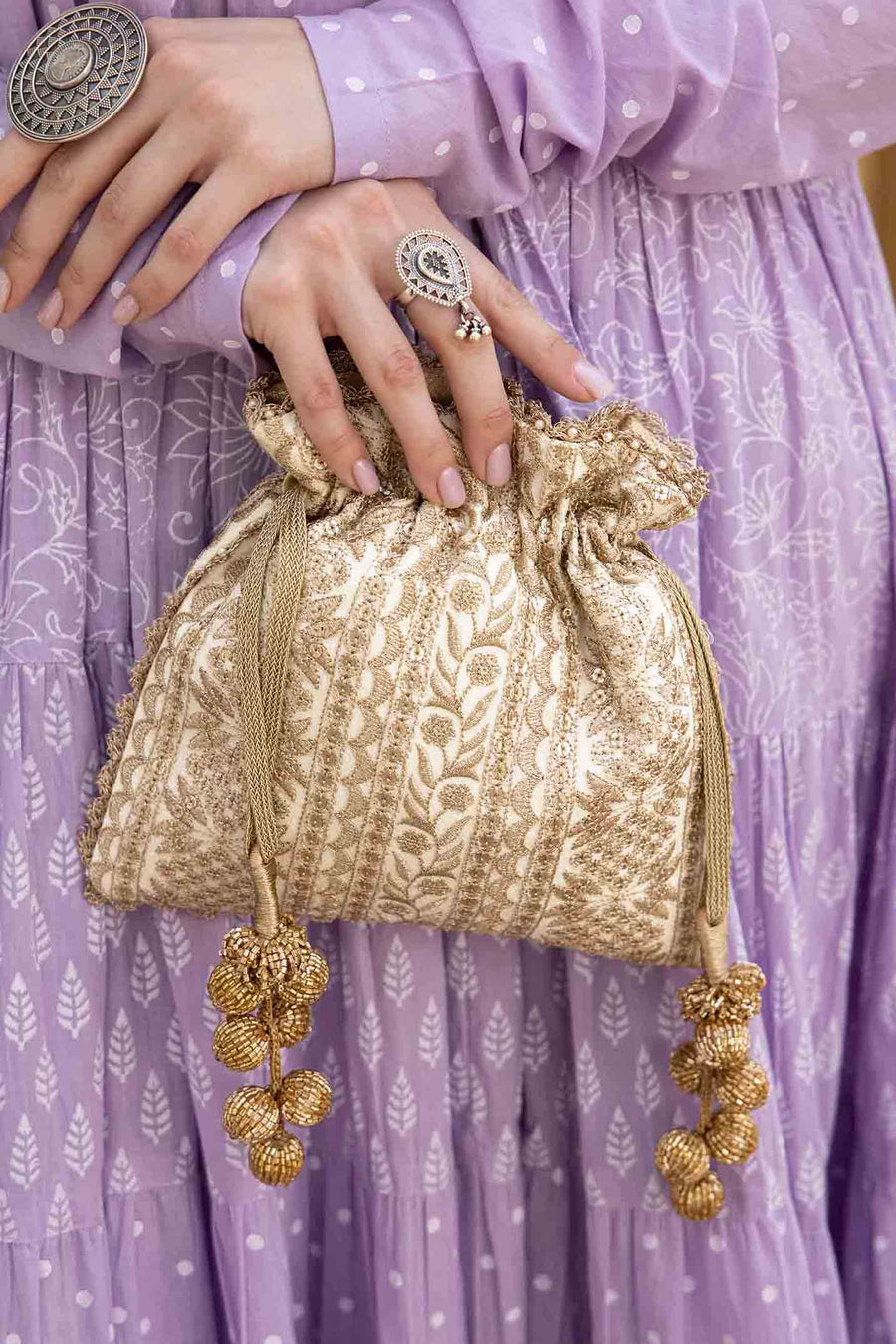 Close-up of a gold embroidered potli bag with drawstring tassels, held by a woman wearing a lavender traditional dress.
