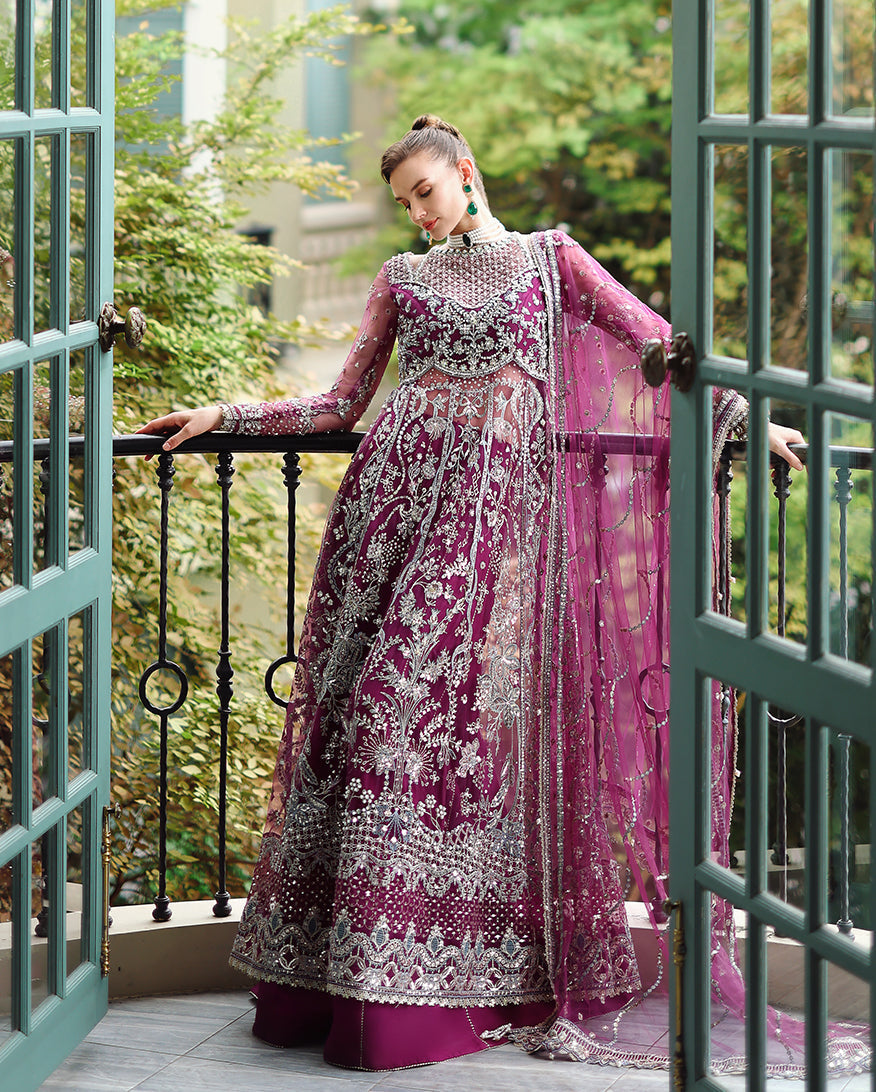 Woman wearing deep magenta formal wedding dress with intricate silver embroidery and sheer long sleeves standing on balcony with green foliage background