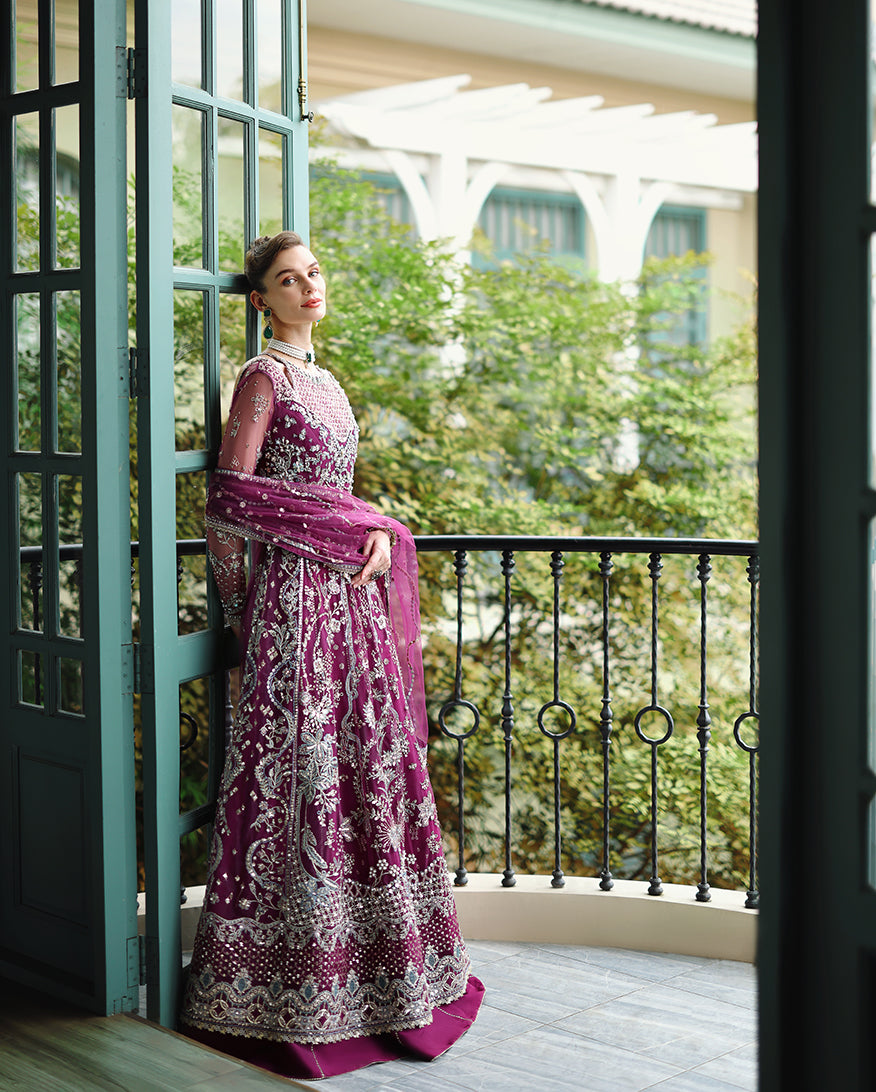 Model in magenta embroidered formal dress with pearl accents and matching sheer dupatta leaning against balcony railing outdoors