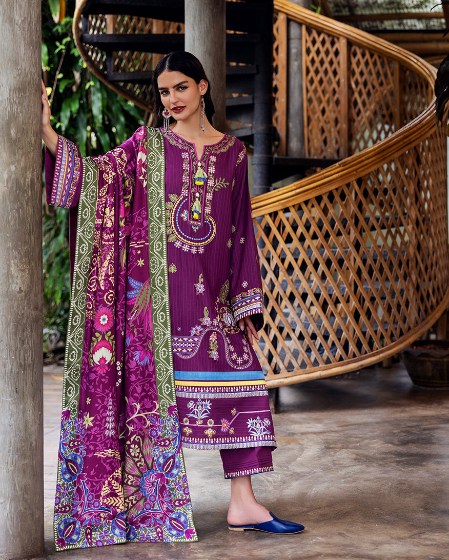 Model in plum purple embroidered Karandi suit standing near a wooden pillar holding matching floral printed shawl.