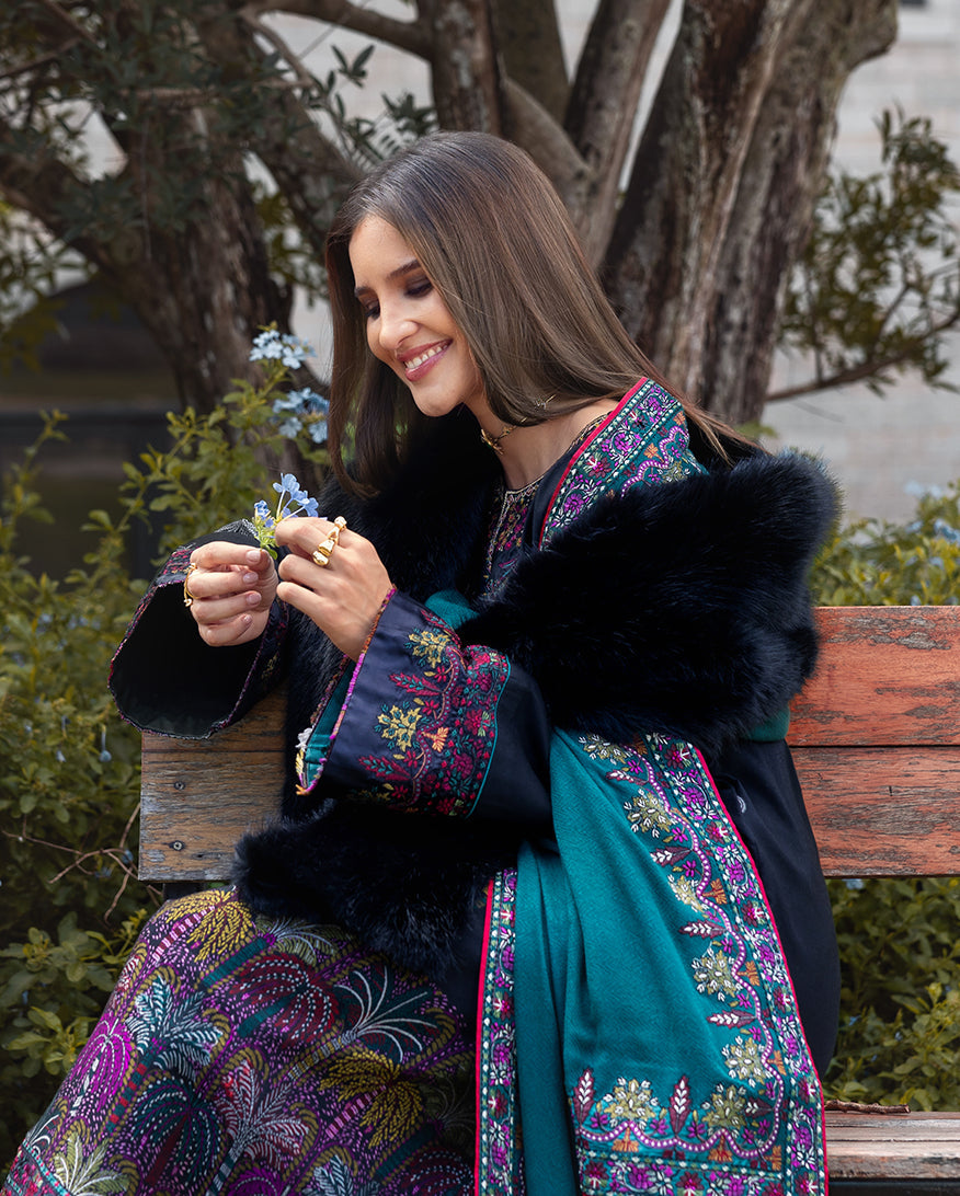 Woman seated on wooden bench wearing navy embroidered winter suit with a black fur shawl over her shoulders