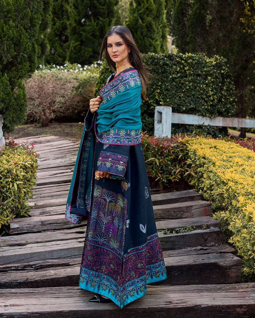 Side view of woman in navy embroidered winter suit and teal shawl standing on wooden pathway surrounded by greenery