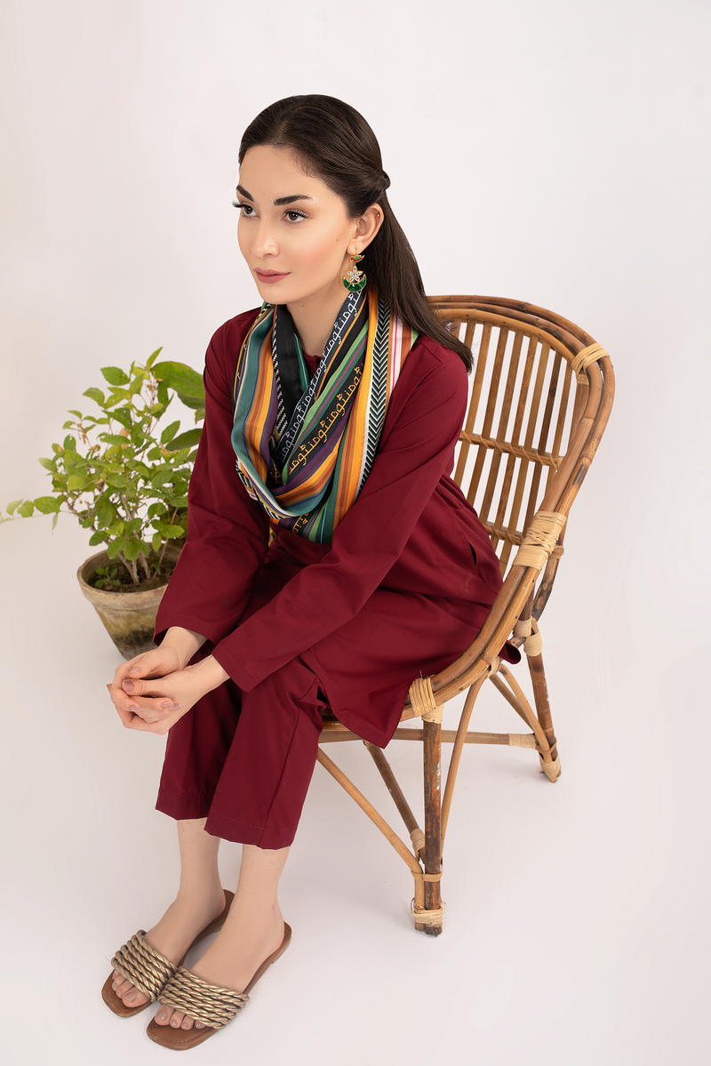 Woman seated in wicker chair wearing a luxurious multicolor striped loop scarf with embroidered script paired with a deep maroon traditional outfit and braided sandals