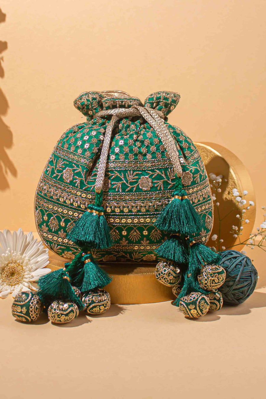Green embroidered potli bag with gold zari work, tassels, and decorative balls, placed on a beige background