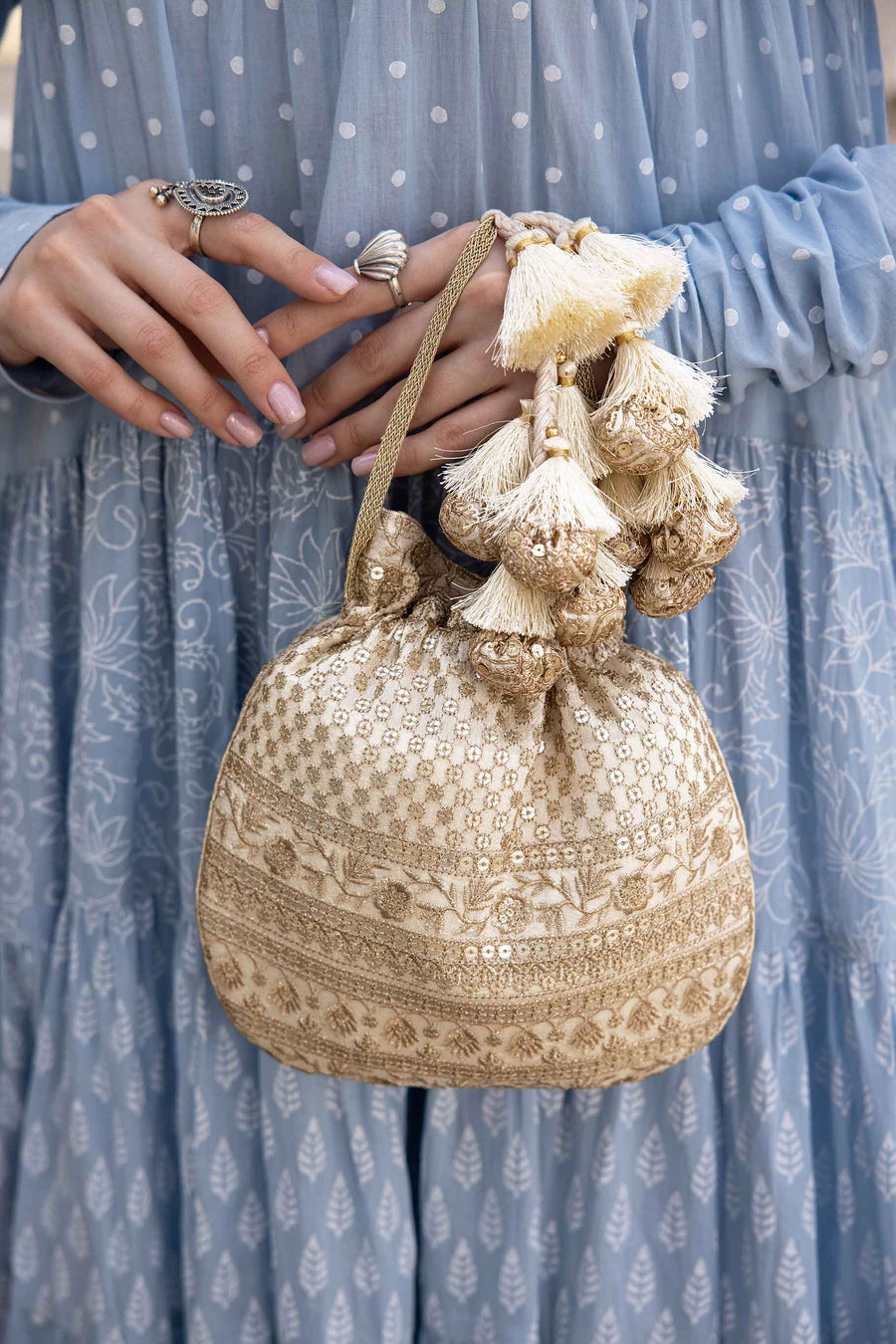 Close-up of off-white potli bag with gold embroidery and tassels held by woman in light blue dress with polka dots
