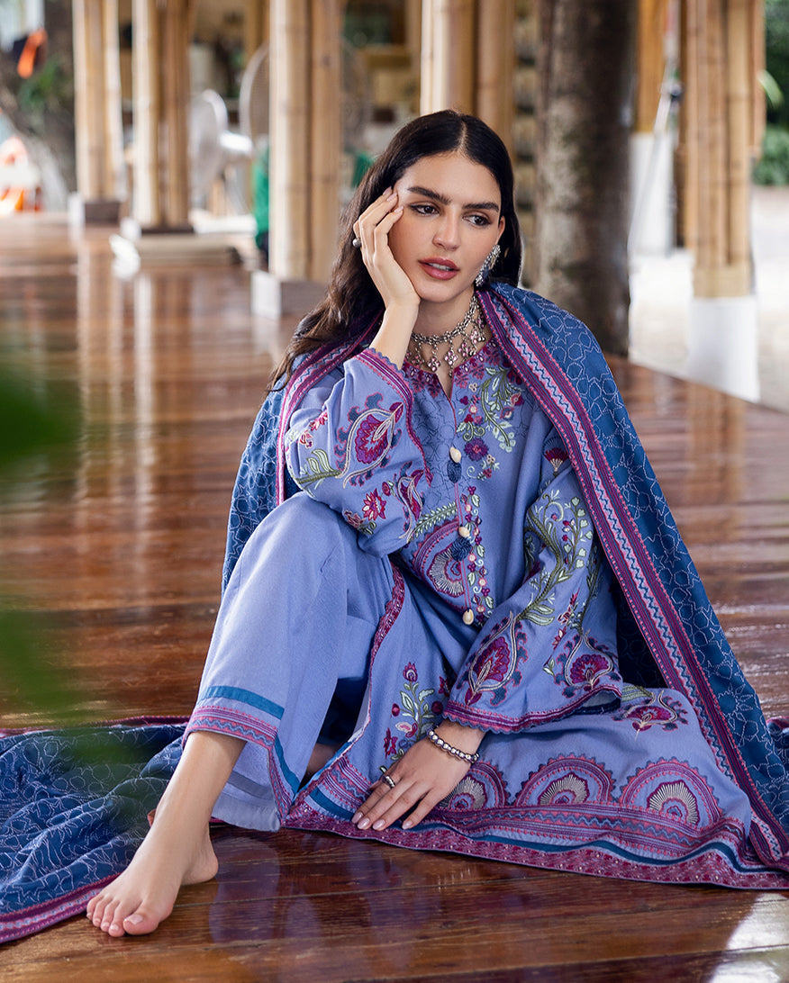 Seated woman in an embroidered lavender Karandi suit with floral designs, wearing a blue shawl and silver jewelry, resting on a wooden floor in a bamboo venue.