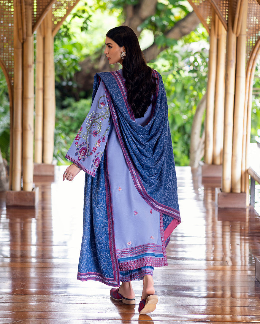 Back view of a woman wearing a lavender embroidered Karandi suit with a deep blue shawl draped over her shoulders, walking along a bamboo pillar-lined wooden corridor.