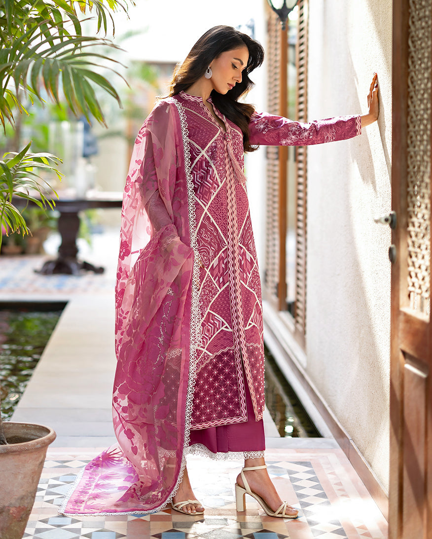 Woman wearing a magenta embroidered three-piece lawn suit standing with hand on wall, showcasing full outfit and sheer floral organza dupatta draped elegantly