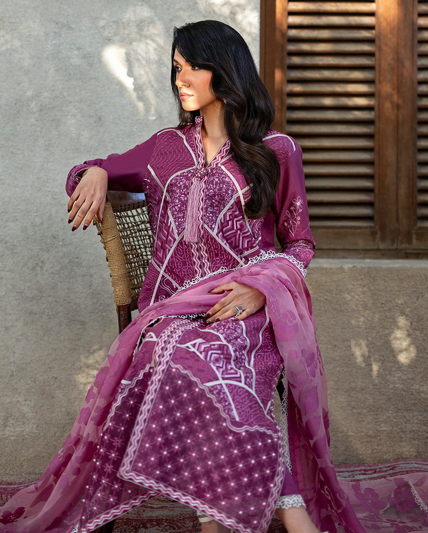 Seated woman in magenta embroidered lawn suit with sheer floral dupatta, showcasing elegance and modest style in a warm, textured setting