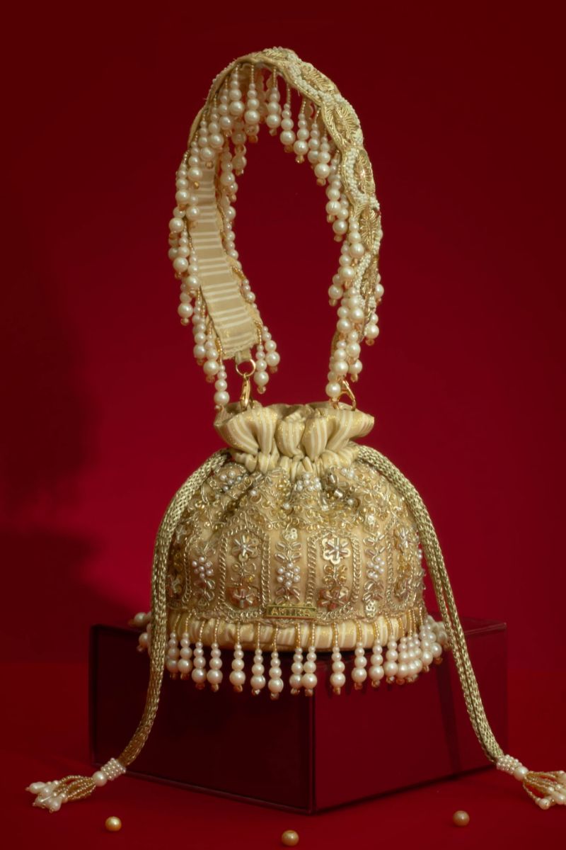 Sajni mini potli bag in gold with detailed pearl embroidery, pearl fringes, and beaded tassel handle displayed on a red background