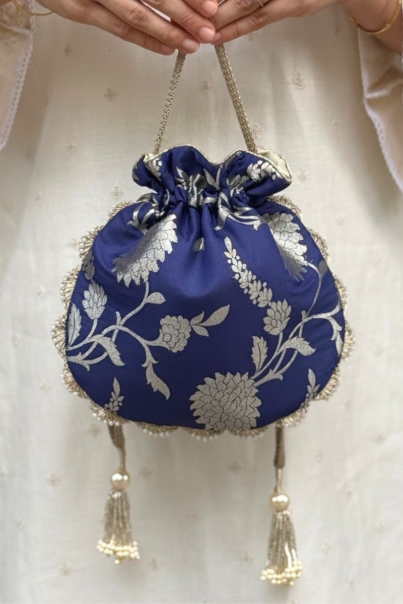 Navy blue Banarasi potli bag with silver floral embroidery and pearl tassels, held by hands in front of cream fabric background