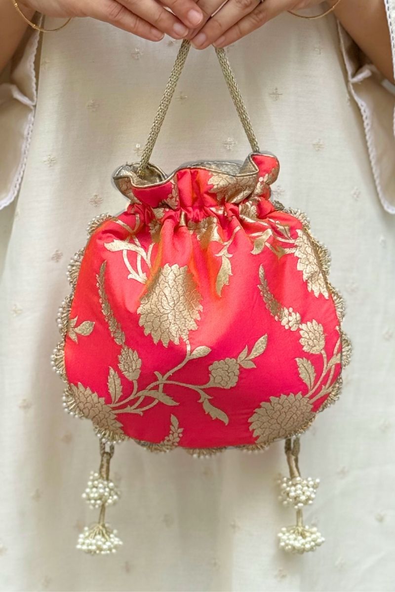 Hand holding coral pink Satrangi Banarasi potli bag with gold floral embroidery and pearl tassels against white embroidered background