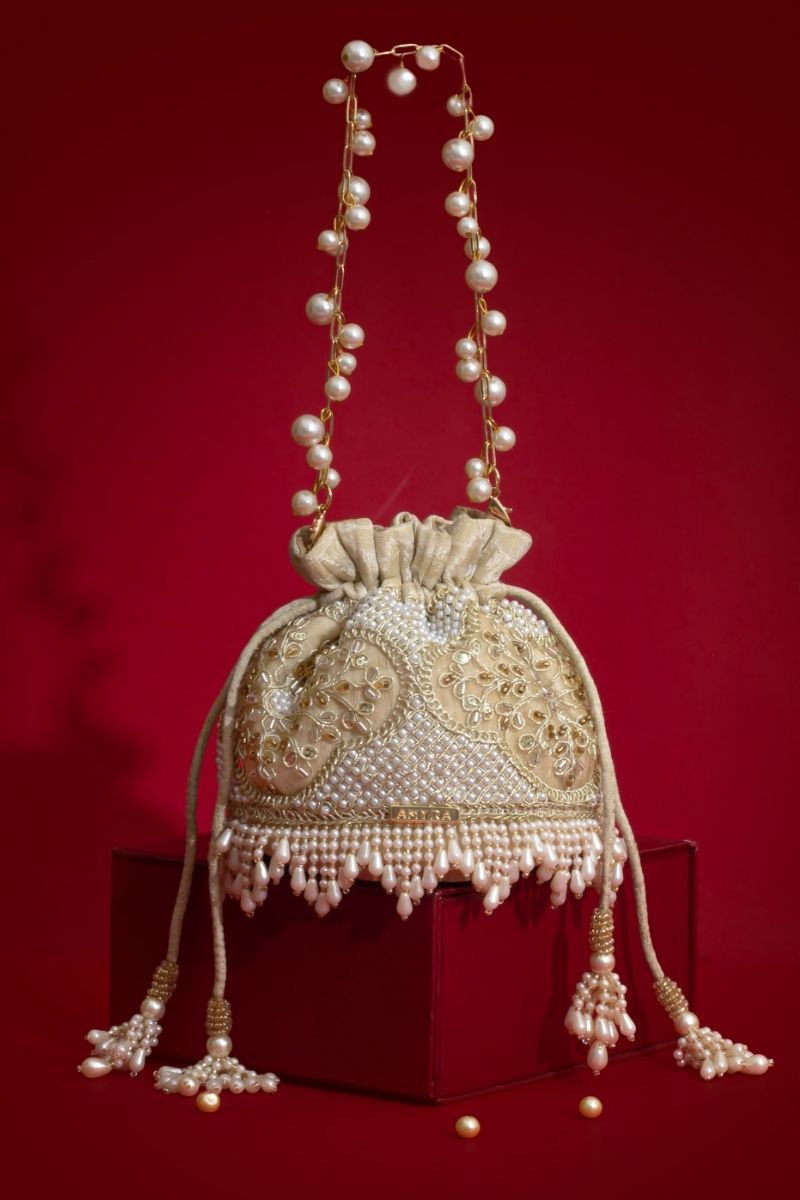Beige Sehra potli bag with intricate pearl embroidery, pearl tassels, and a pearl-adorned handle displayed against a red background.