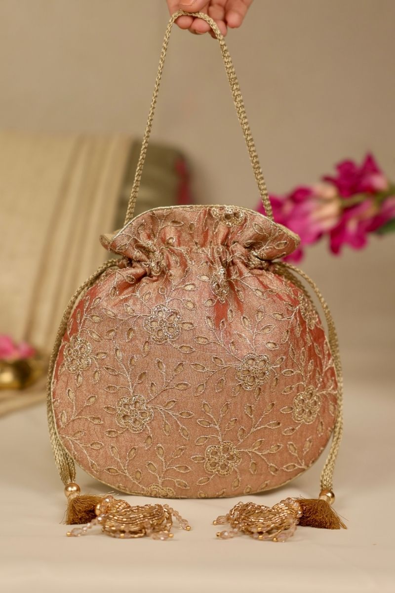 Hand holding embroidered copper potli bag with golden floral embroidery and decorative tassels against a neutral background