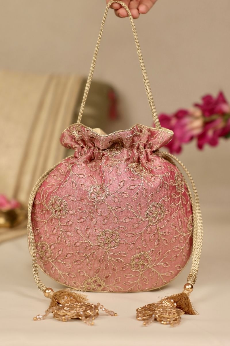 Pink potli bag with intricate gold floral embroidery, drawstring closure, and tasseled ends, held by a braided handle.