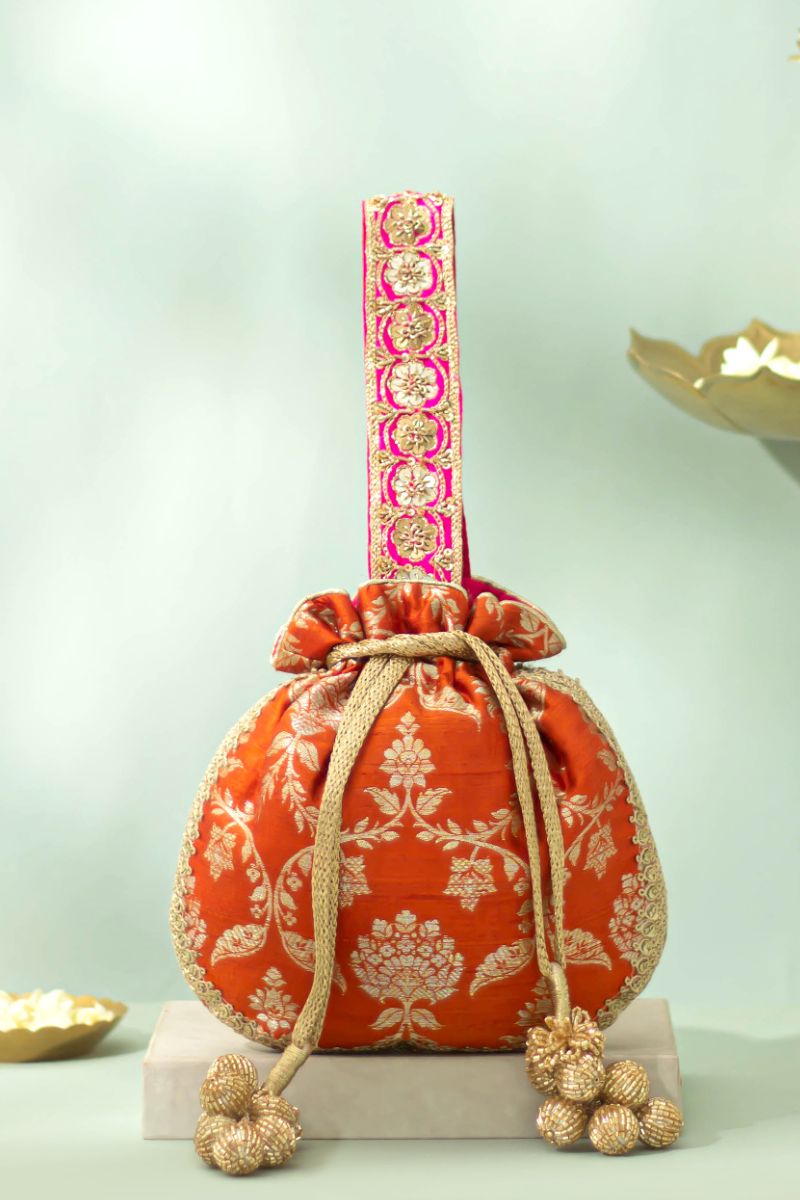 Rust orange pure raw silk potli bag with gold floral embroidery, embellished pink handle with gold zardosi flowers, lace trim, and gold cutdana tassels