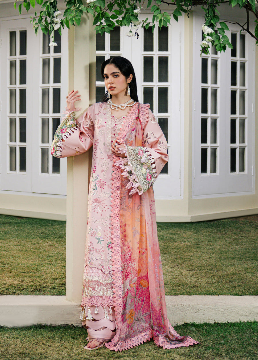 Woman wearing a baby pink embroidered luxury lawn suit with floral designs and lace trims, standing outdoors near a building with white windows.