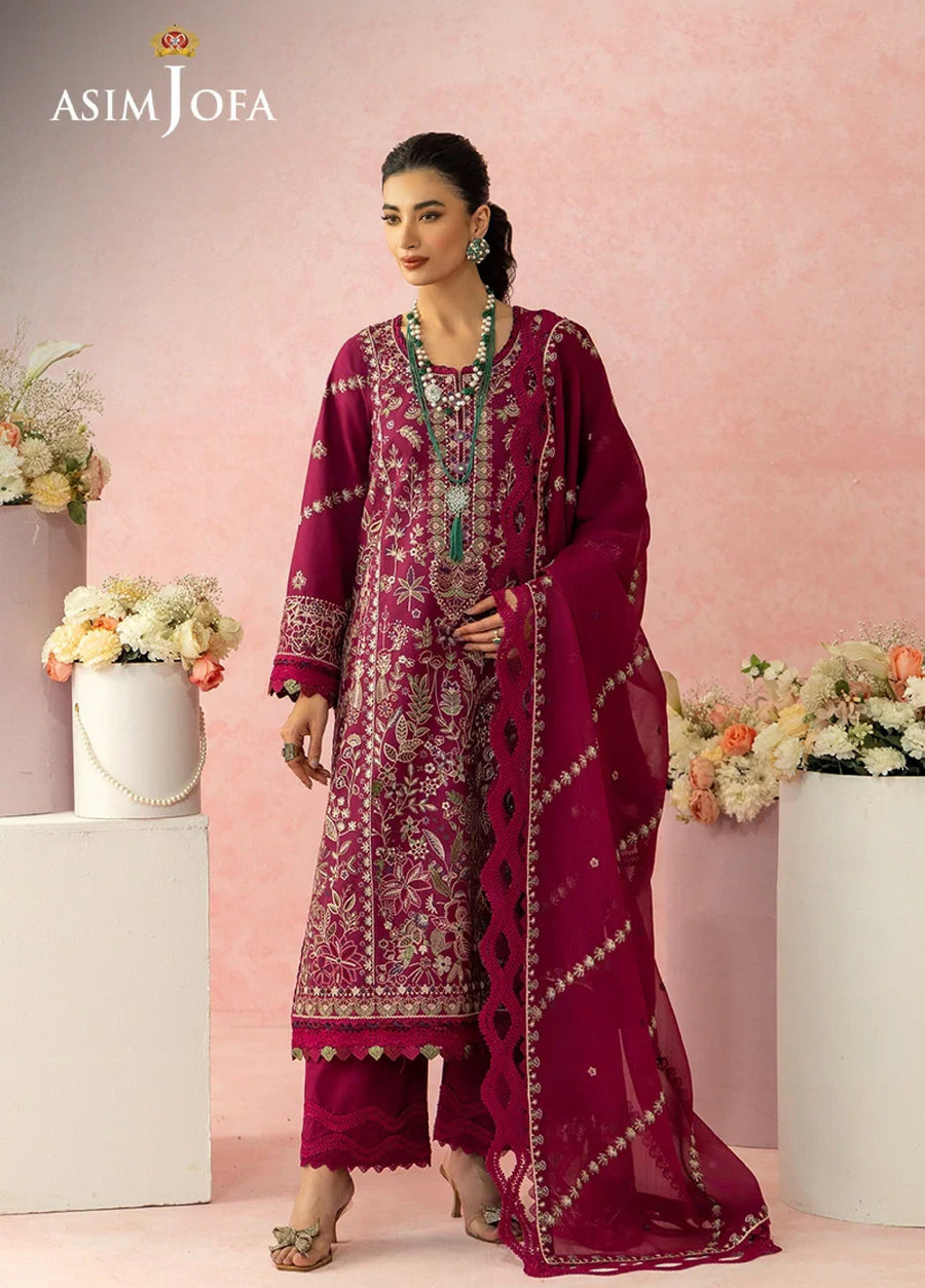 Woman modeling magenta embroidered luxury lawn suit with floral motifs and scalloped edges holding a chiffon dupatta, standing against a pink backdrop with floral decor.
