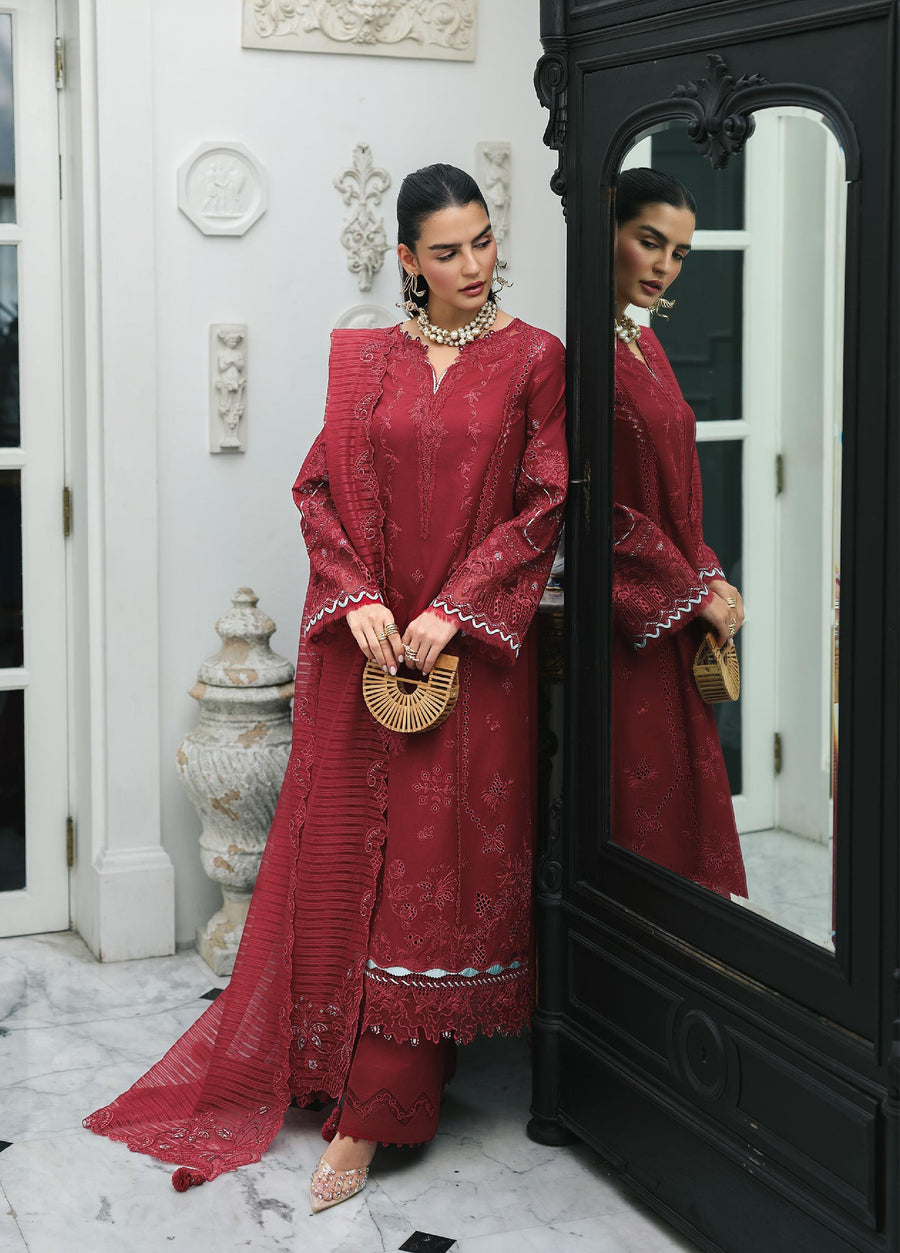 Woman wearing a maroon embroidered lawn suit with intricate threadwork, holding a wooden handbag, standing beside a mirror in a formal indoor setting.