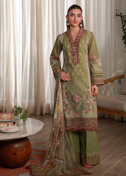 Front view of a woman wearing an olive green embroidered lawn suit with pink and beige floral designs and a printed chiffon dupatta, standing in a softly lit room.