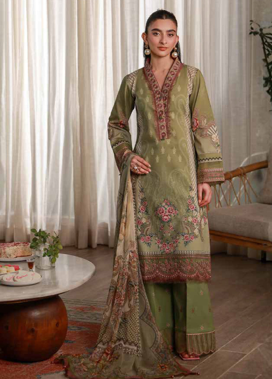 Front view of a woman wearing an olive green embroidered lawn suit with pink and beige floral designs and a printed chiffon dupatta, standing in a softly lit room.