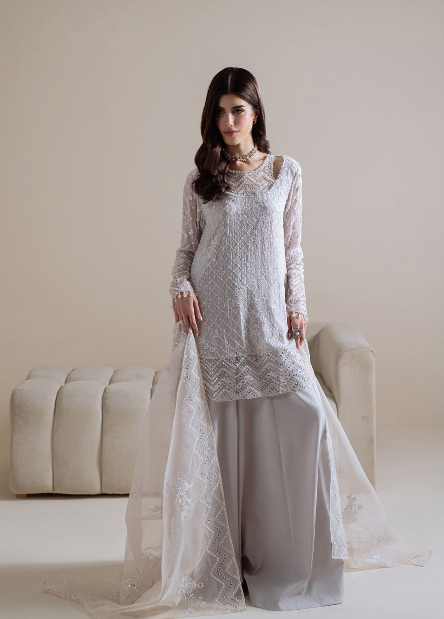 Model wearing a full white embroidered chiffon tunic paired with light gray silk trousers and an embroidered sheer dupatta in an elegant formal setting