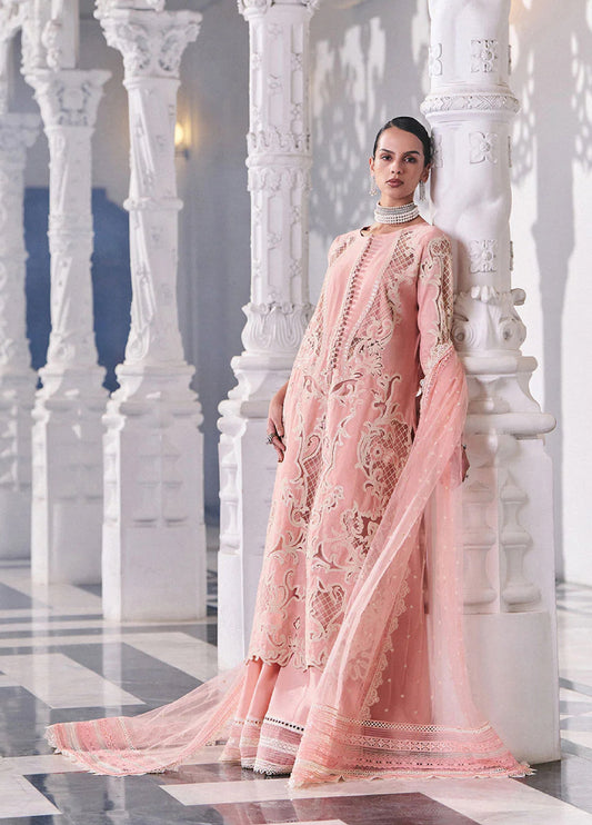 Model wearing an elegant soft pink embroidered lawn suit with intricate lace and embroidery, standing among white carved pillars in a luxurious hall.