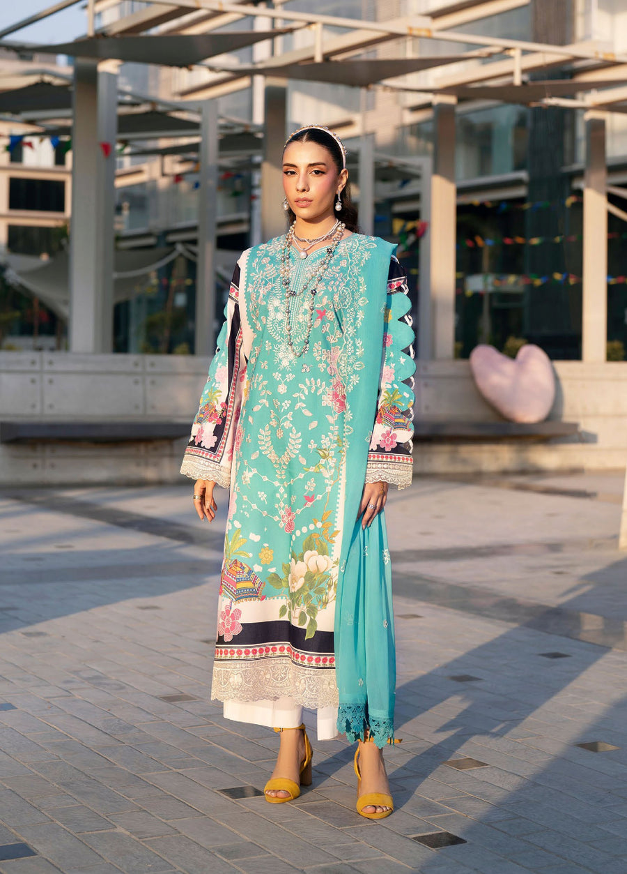Front view of woman wearing turquoise embroidered lawn suit with floral patterns, paired with white trousers and yellow heels.