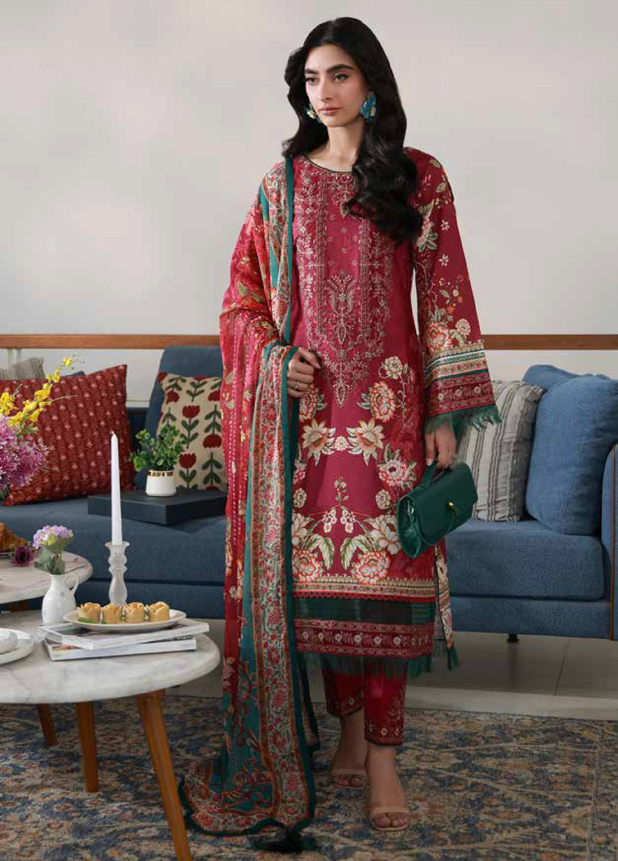 Woman in a rich red embroidered floral lawn suit holding a green handbag, standing in a living room with blue and patterned cushions, showcasing the front of the outfit with detailed embroidery and printed chiffon dupatta