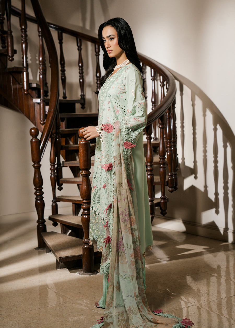 Side profile of a woman wearing a pastel mint green embroidered lawn suit with a floral chiffon dupatta standing beside a wooden spiral staircase.