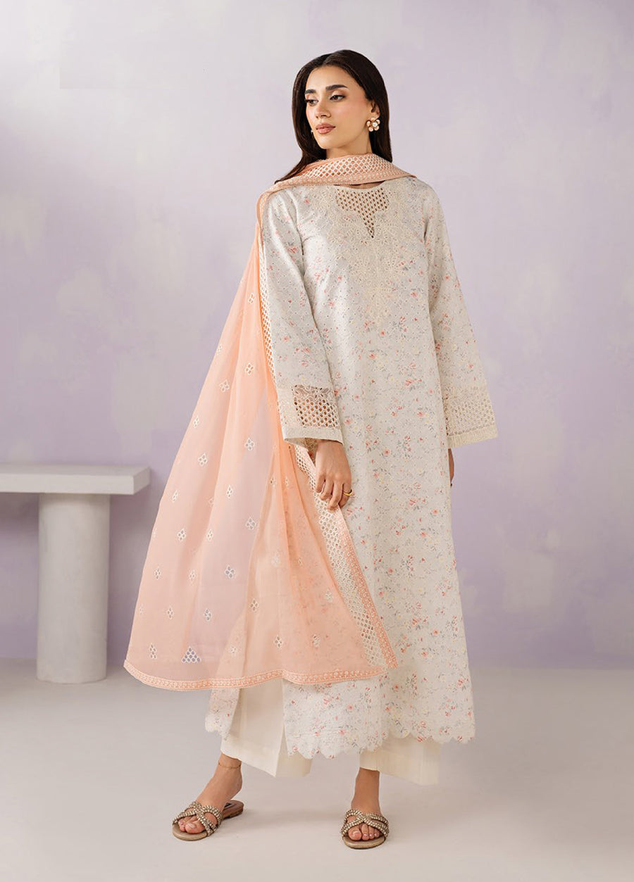 Woman wearing an embroidered floral lawn suit in light beige with peach chiffon dupatta and white trousers standing against pastel background