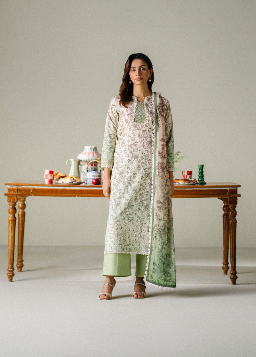 Woman wearing an elegant cream and pastel green printed lawn suit with embroidered neckline and lace borders, standing in front of a wooden table with decorative items.