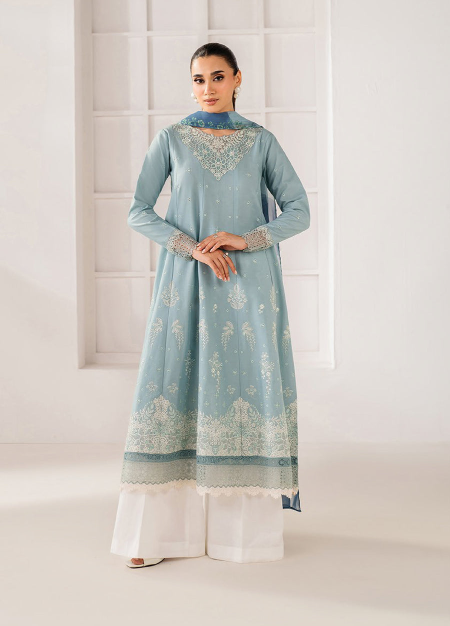 Woman wearing a sky blue embroidered lawn suit with lace detailing on neckline and sleeves, paired with white trousers in a minimalist indoor setting.