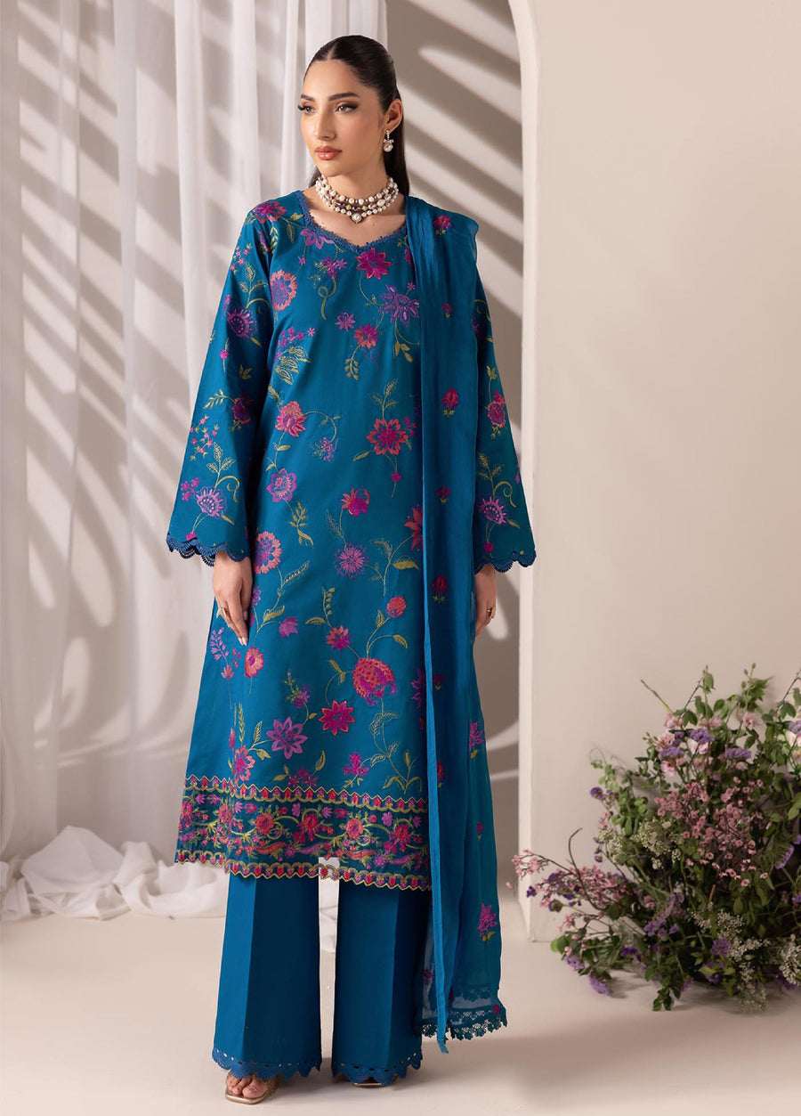 Woman wearing a deep blue embroidered lawn shirt with vibrant pink floral patterns, matching blue wide-leg pants, and coordinating chiffon dupatta in a soft-lit indoor setting