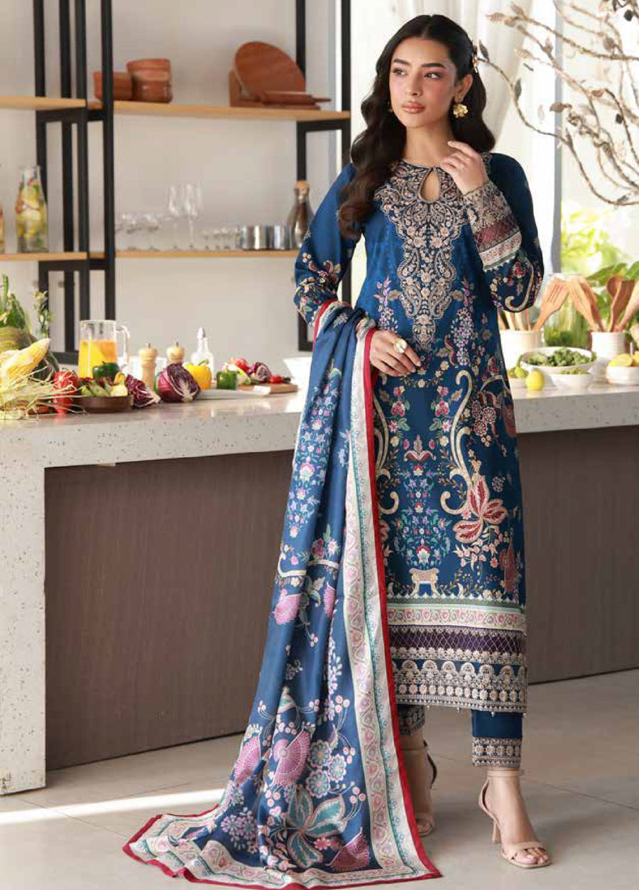 Front view of a woman wearing a deep blue embroidered lawn suit with floral and paisley patterns, holding a matching printed chiffon dupatta in a modern kitchen setting