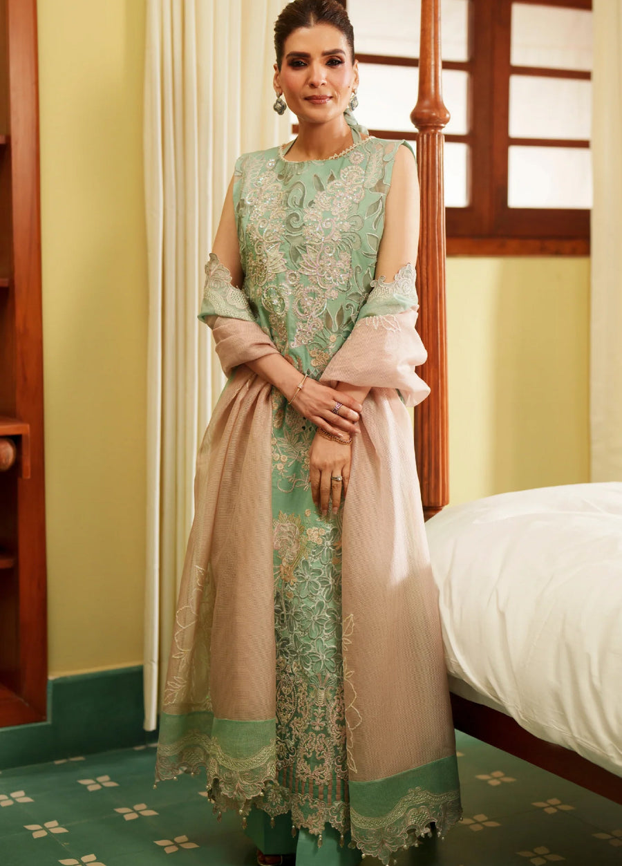 Woman wearing a sleeveless green embroidered lawn suit with intricate floral designs and a beige embroidered shawl, standing indoors.