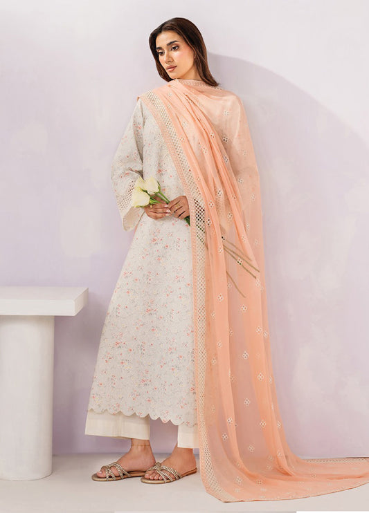 Side view of woman holding white roses wearing embroidered floral beige lawn suit with scalloped hem, sheer peach dupatta with embroidery, and white trousers