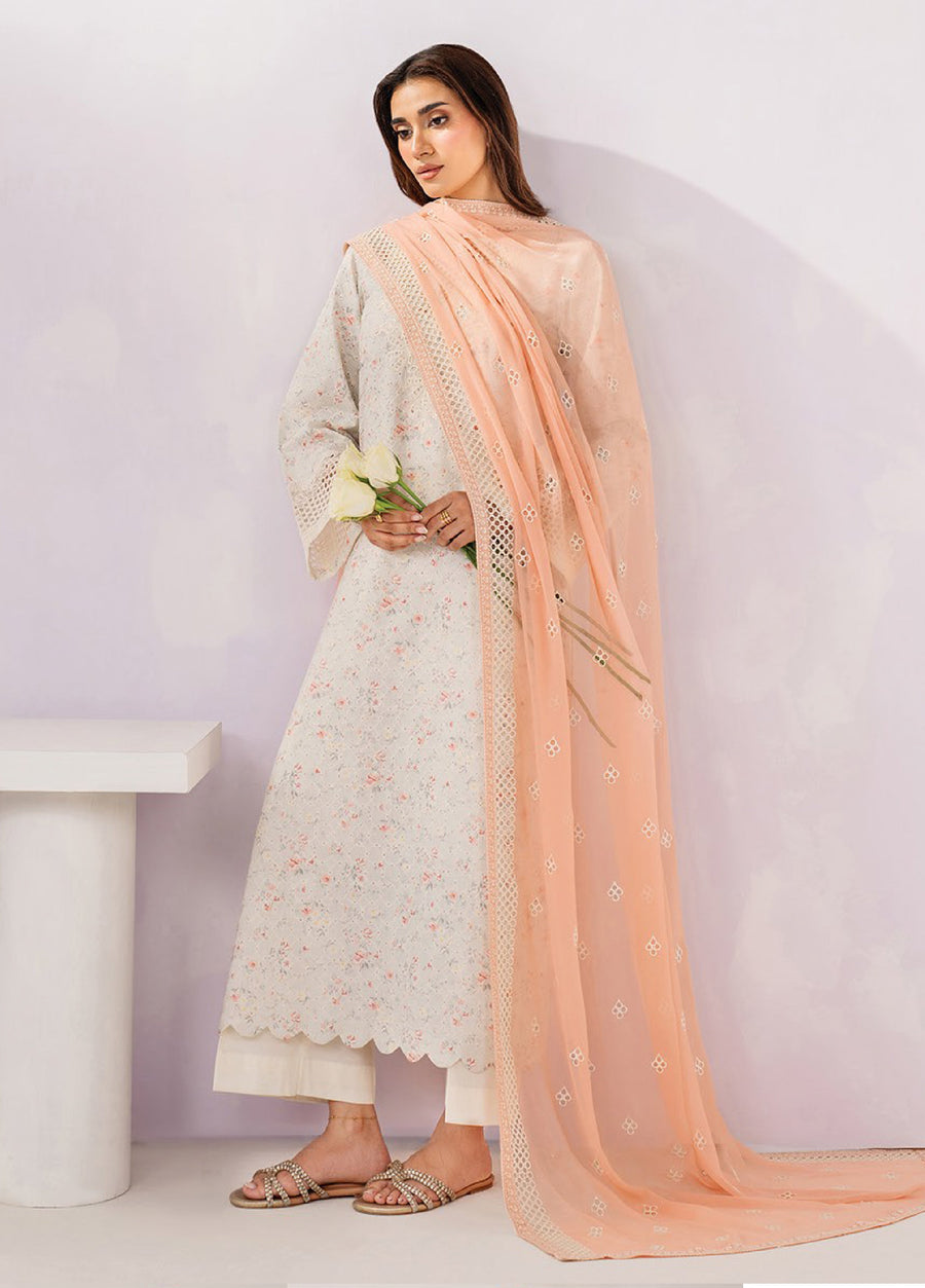 Side view of woman holding white roses wearing embroidered floral beige lawn suit with scalloped hem, sheer peach dupatta with embroidery, and white trousers