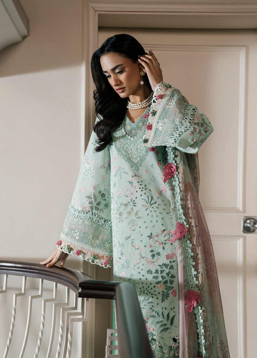 Close-up of a woman in a pastel mint green lawn suit with detailed floral embroidery and lace on the sleeves, holding a floral chiffon dupatta near a staircase railing.