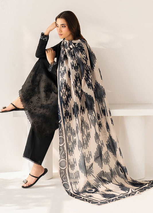 Side view of a woman seated, showcasing the intricate black embroidery on her black formal suit and the flowing beige dupatta with bold black geometric prints.