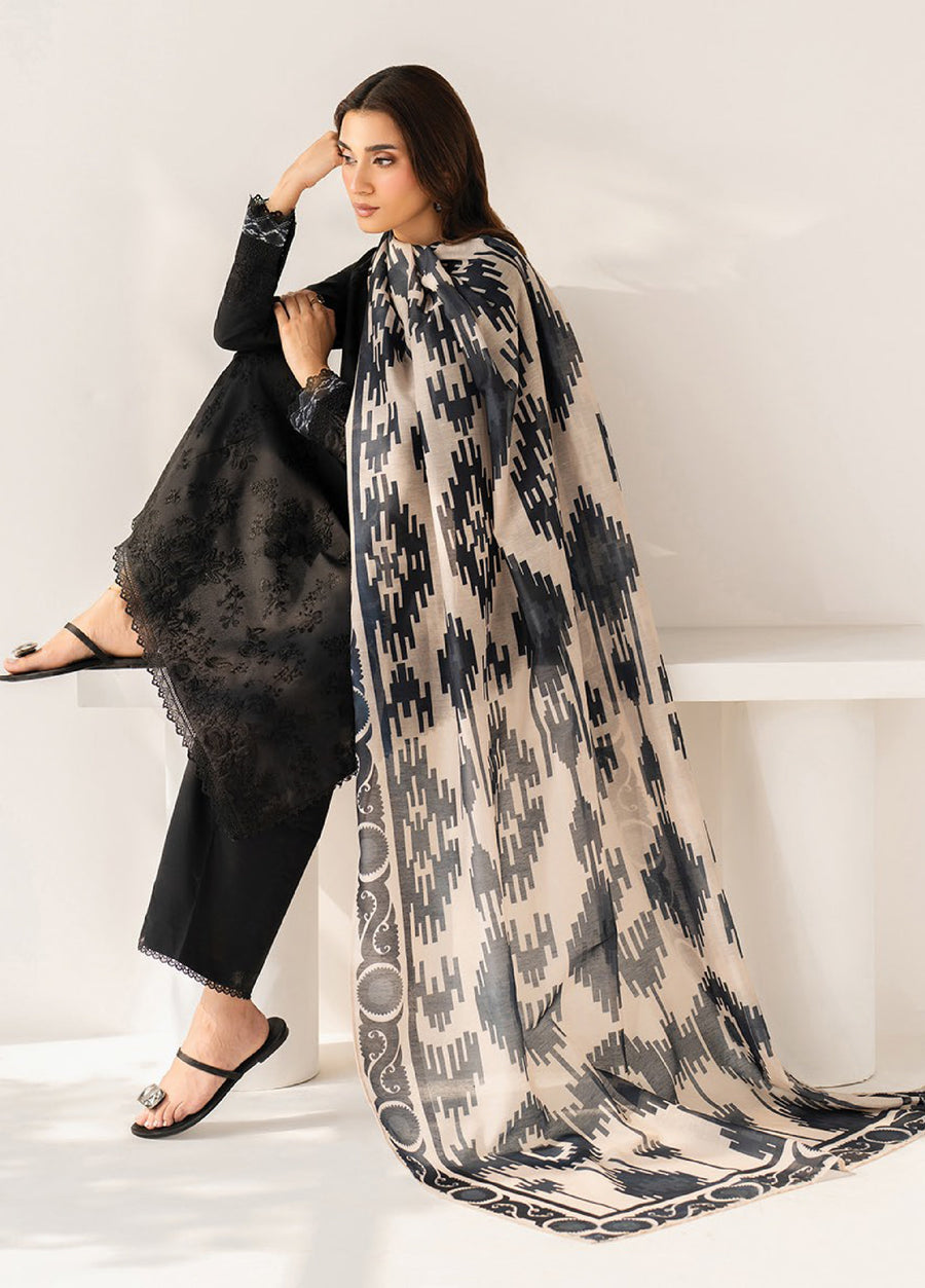 Side view of a woman seated, showcasing the intricate black embroidery on her black formal suit and the flowing beige dupatta with bold black geometric prints.