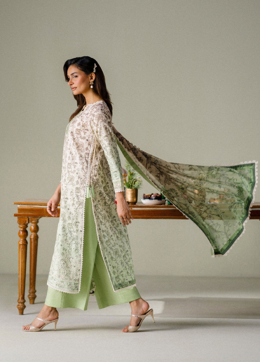 Side view of woman walking gracefully in a floral-printed lawn shirt with chiffon dupatta flowing behind her and light green cambric trousers.