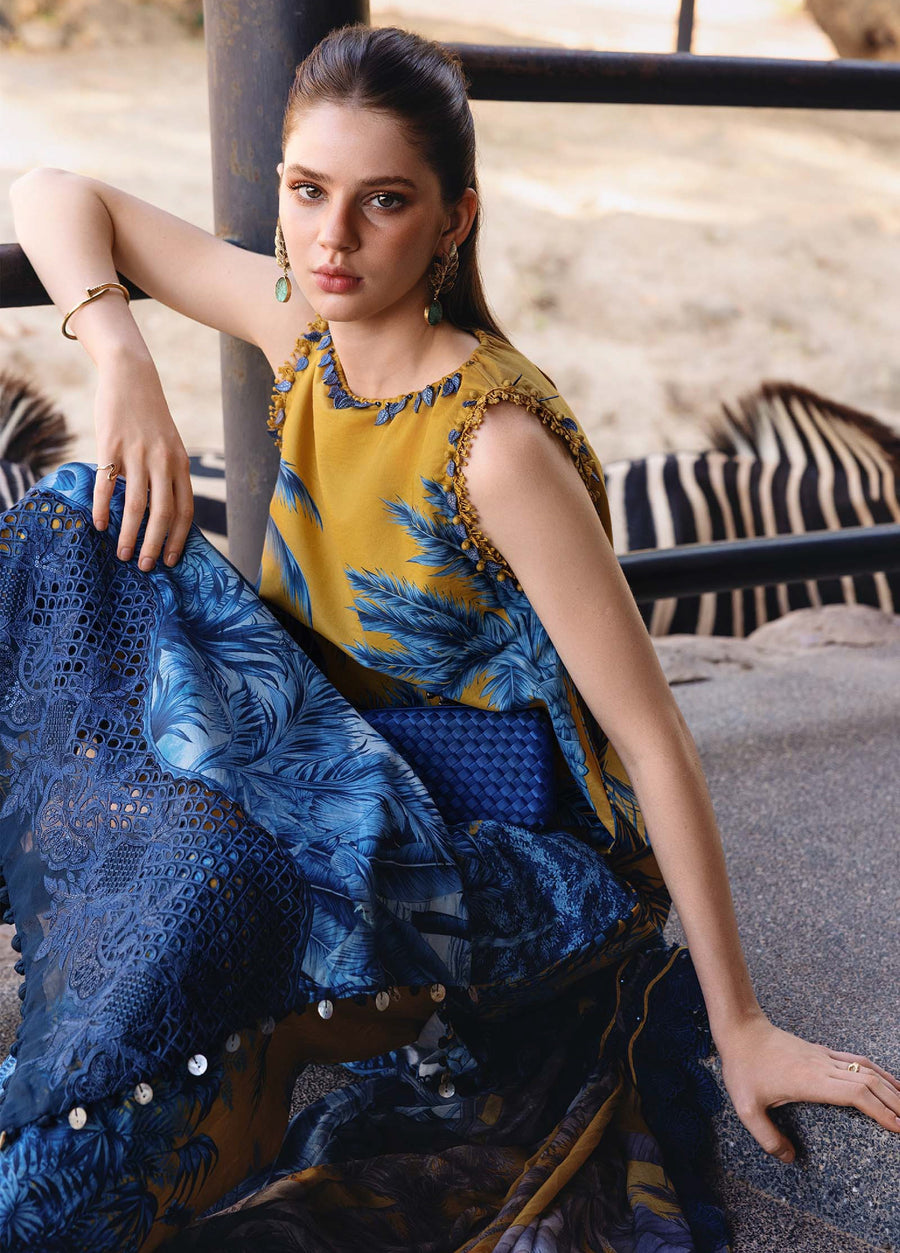 Close-up of a woman in a mustard yellow and blue printed lawn suit showcasing embroidered sleeve trim and holding a blue clutch