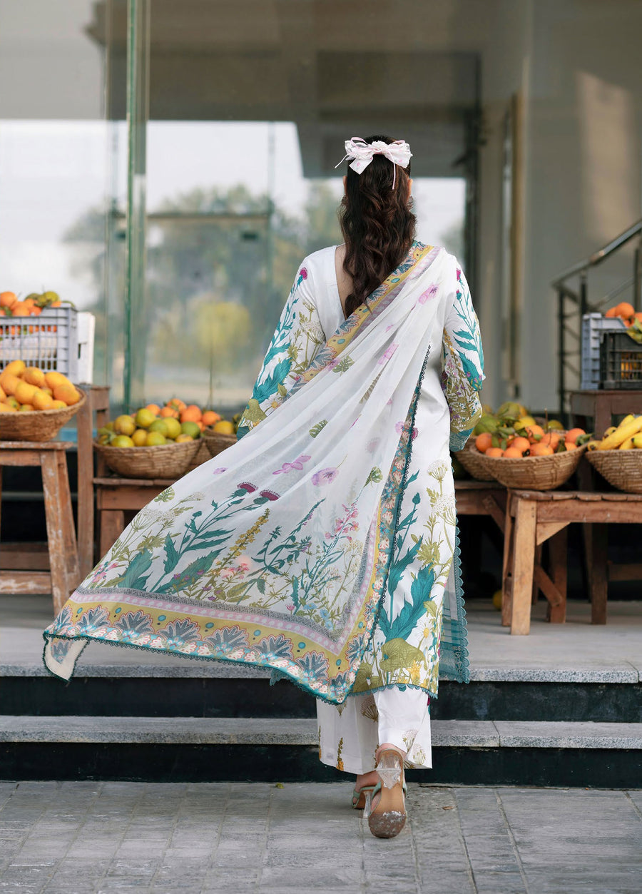 Back view of woman in white and teal embroidered lawn suit with floral chiffon dupatta flowing, standing outdoors with fruit baskets behind her.