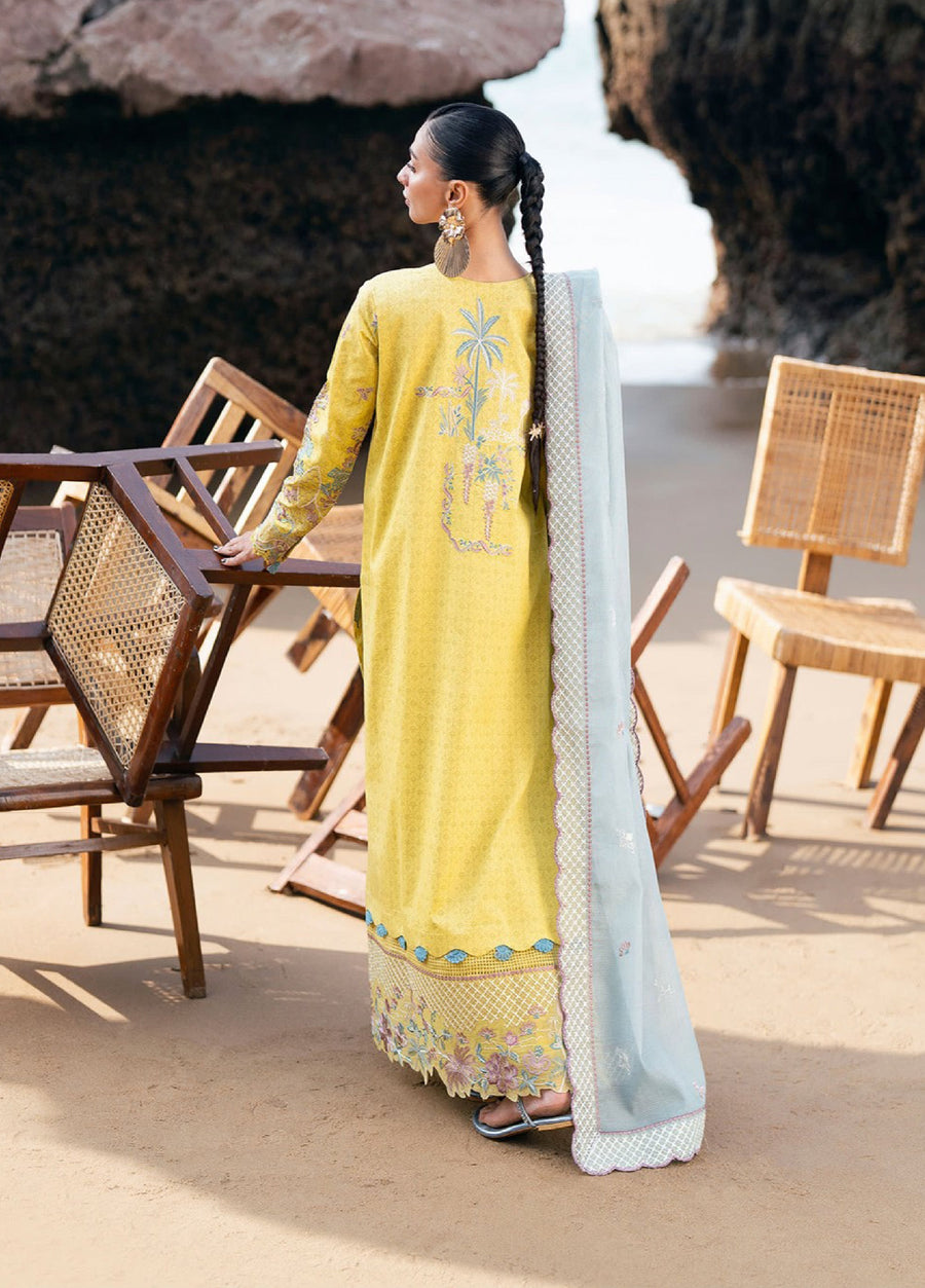 Rear view of woman wearing the yellow embroidered lawn suit showing detailed embroidery on the back and the light blue dupatta draped over one shoulder, standing next to wooden chairs on sand near rocks.